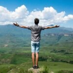 A man stands triumphantly with arms open, overlooking a scenic mountain range.