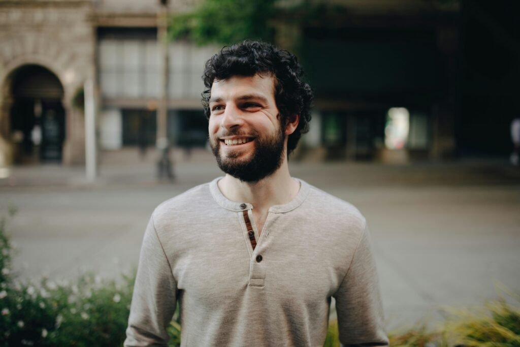 pexels-photo-8575396-8575396 A bearded man with curly hair smiling outdoors in a casual setting.